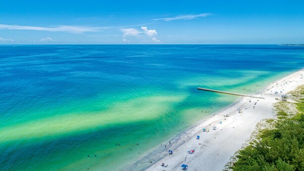 A beach with clear turquoise waters, a pier extending into the sea, and a sandy shore with umbrellas and beachgoers. - Cottage Haven-one Minute Walk To The Beach-private Yards-keyless Locks (Bradenton Beach)