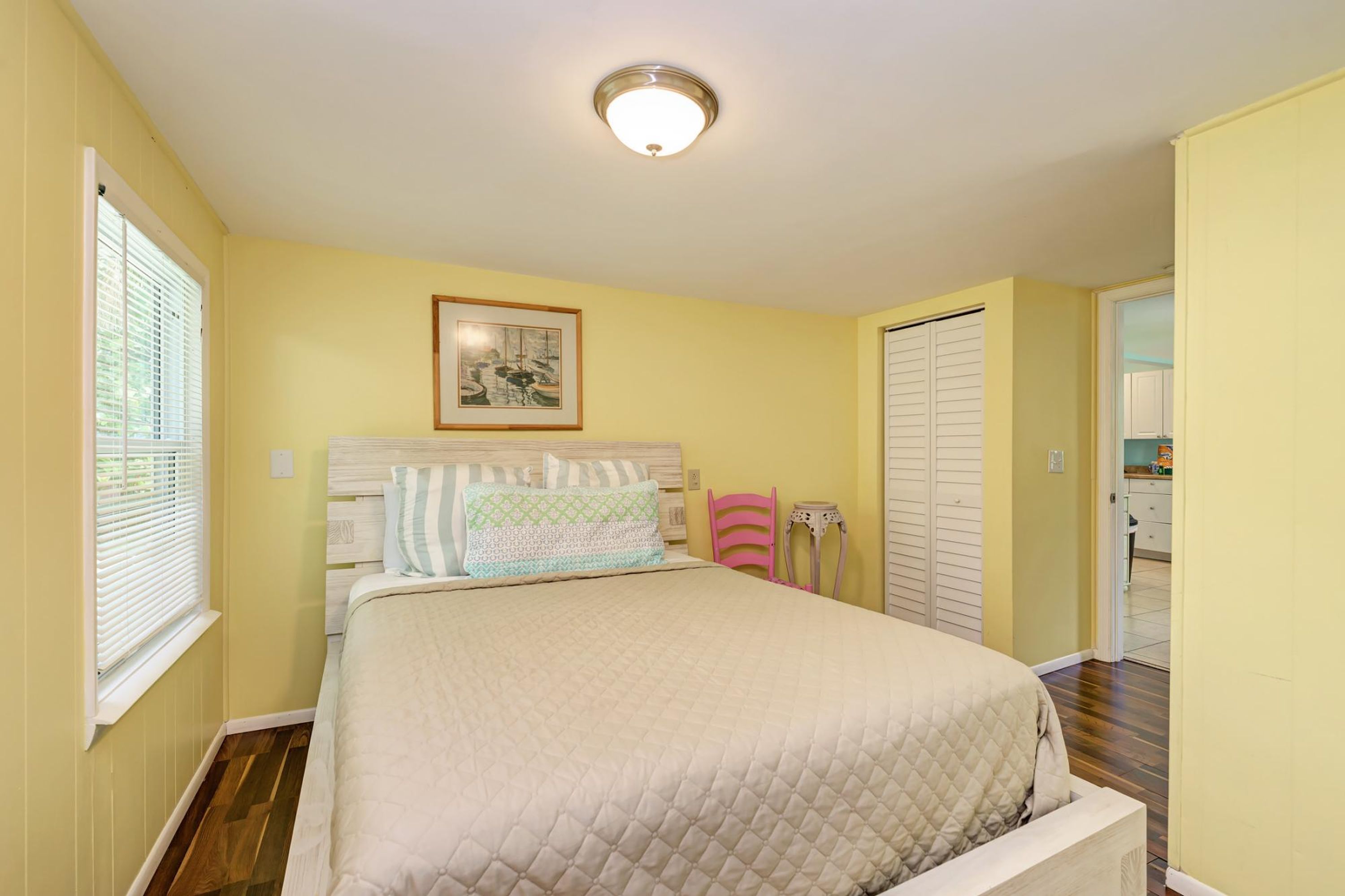 A bedroom with a bed, a bedside table, a chair, a window with blinds, and a door leading to another room.