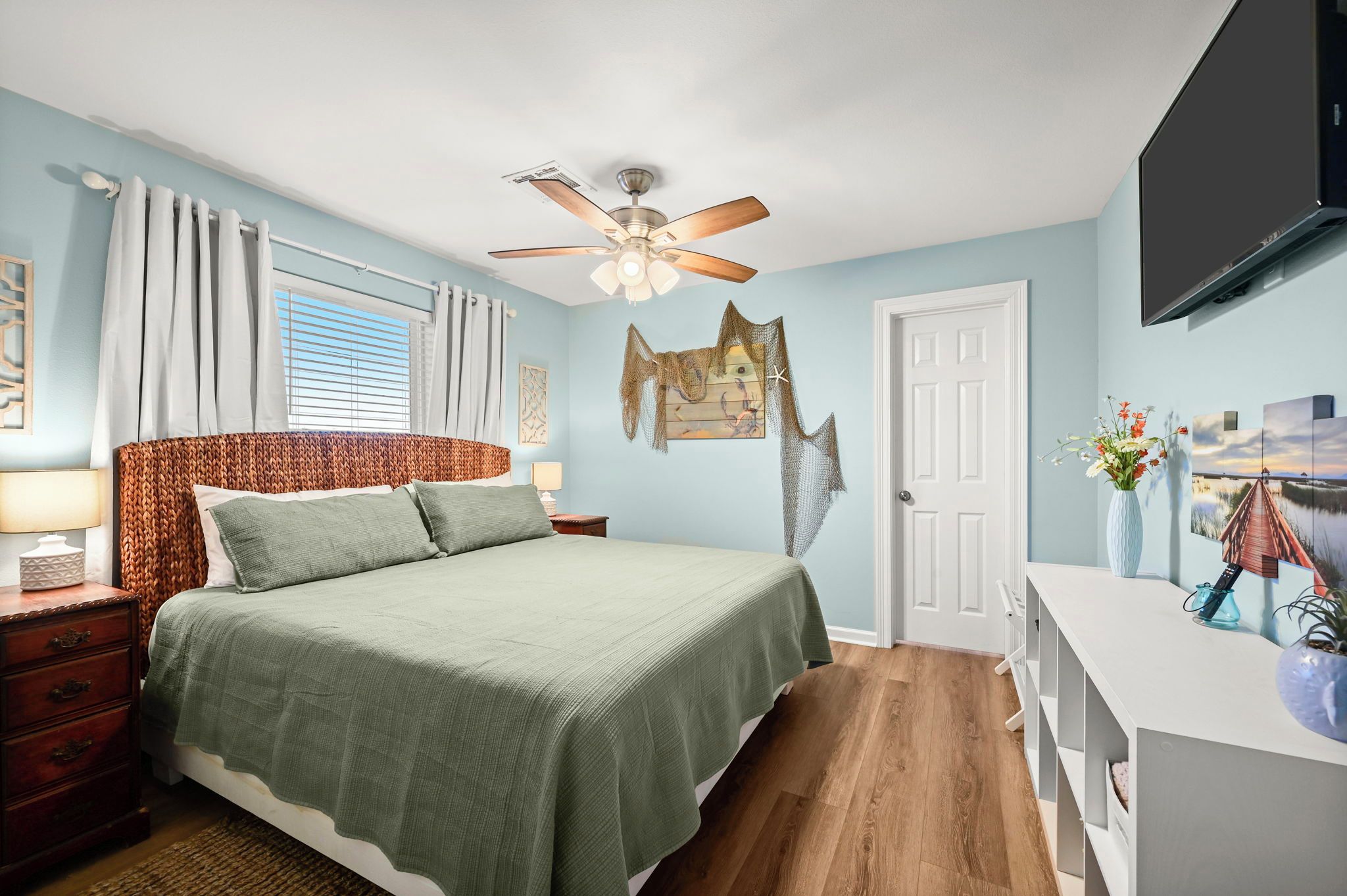 A bedroom with a bed, a nightstand, a dresser, a ceiling fan, a door, and artwork on the walls.