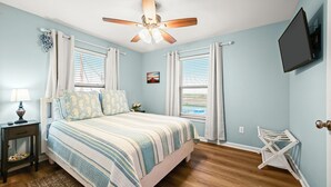 A bedroom with a bed, a ceiling fan, a TV mounted on the wall, and a window with blinds.