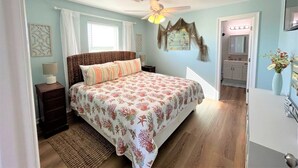 Une chambre à coucher comprenant un lit, des tables de chevet, un ventilateur de plafond, une fenêtre avec des rideaux, une œuvre d’art accrochée au mur et une salle de bain visible à travers une porte ouverte.