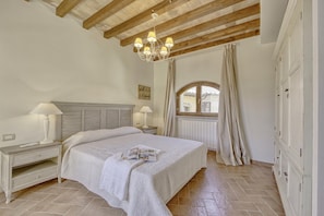 Ein Schlafzimmer mit Bett, Nachttischen, einer Chandelierleuchte, einem Fenster mit Vorhängen und einem Kleiderschrank.