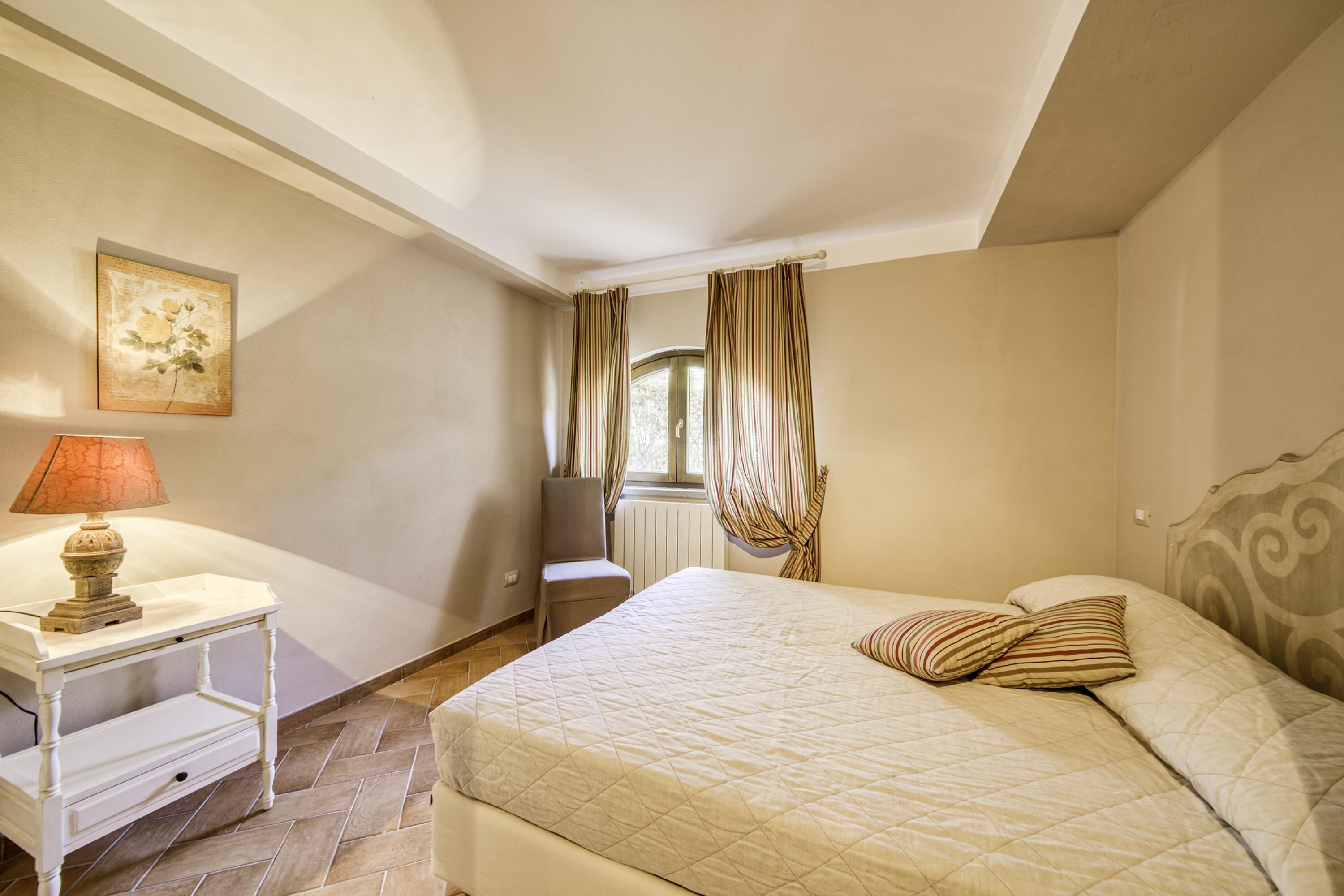 A bedroom with a bed, a nightstand, a lamp, a chair, a window with curtains, and a painting on the wall.