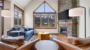 Un salon doté d’une cheminée en pierre, d’une grande fenêtre offrant une vue sur les montagnes, d’un canapé bleu et de fauteuils en cuir.