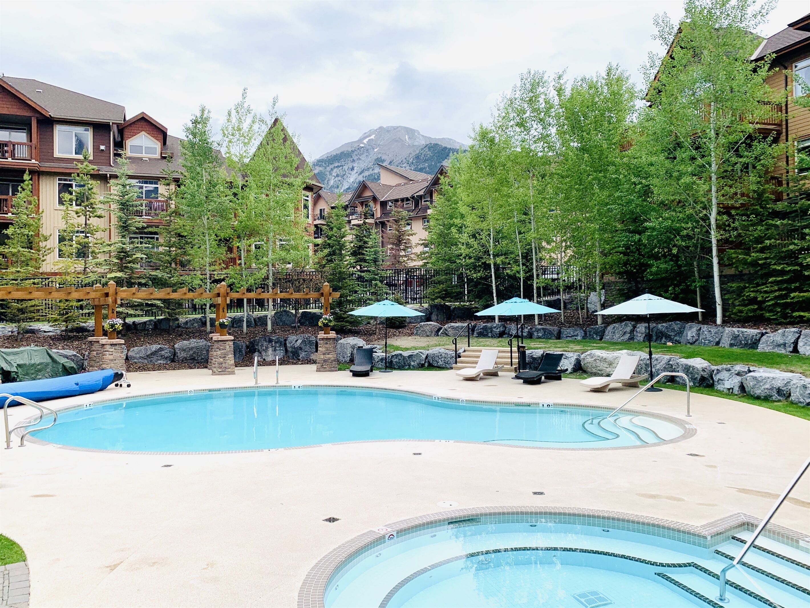 Una piscina con lettini e ombrelloni, circondata da alberi e edifici con montagne sullo sfondo.