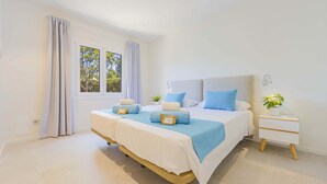 Una cama doble con colcha blanca y azul, dos almohadas, una mesita de noche con lámpara y una ventana con cortinas.