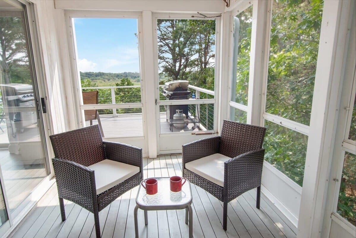 A covered outdoor seating area with wicker chairs and a small table, overlooking a deck with outdoor furniture and a scenic view.