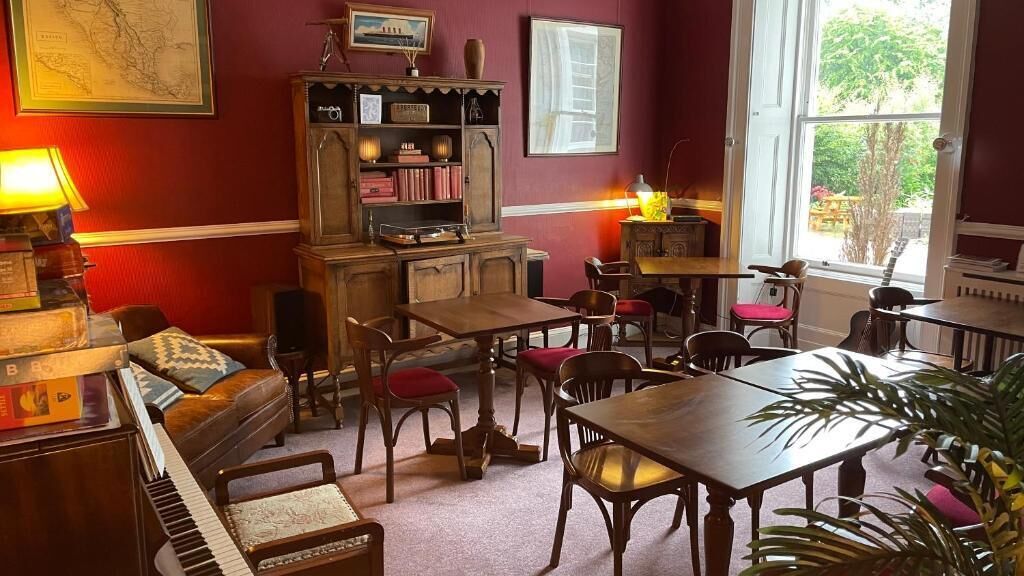 Een kamer met een piano, een bank, een houten tafel en stoelen. Een boekenkast met boeken en decoratieve objecten. Een raam zorgt voor natuurlijk licht.