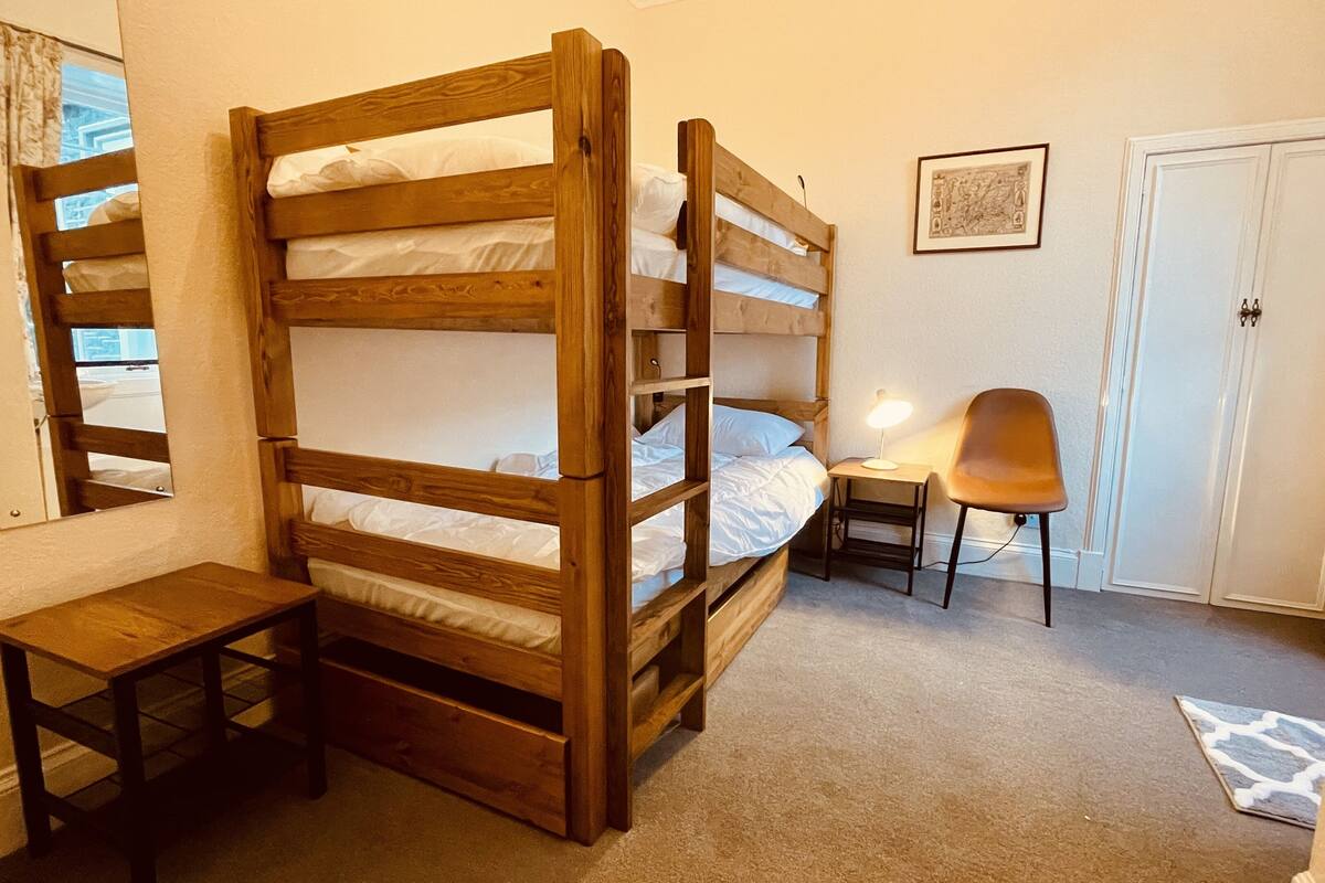 Un lit superposé avec un bureau et une chaise, une petite table et une porte, le tout dans une pièce dont le sol est recouvert d’une moquette.