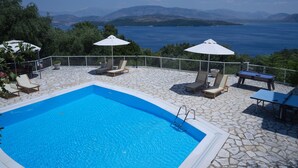A swimming pool with lounge chairs and umbrellas, surrounded by stone flooring and a view of the sea and mountains. - Bellina Villa, Corfu (Αγνίτσινη)