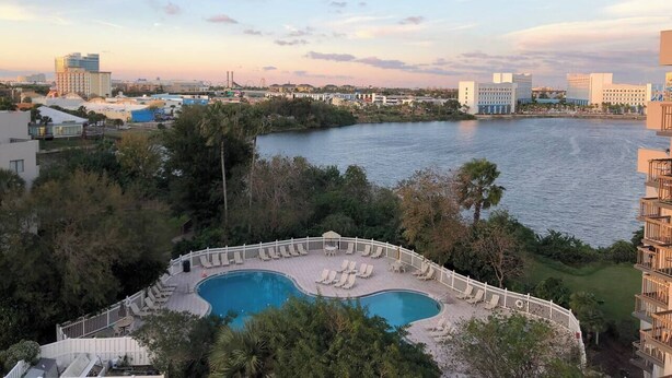 A resort with a large pool surrounded by a white fence, overlooking a body of water with buildings in the background. - Walk to Universal Indoor/outdoor Pool Remodeled! (Orlando)