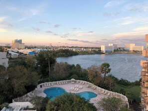Un resort con una piscina grande rodeada por una valla blanca, con vistas a una masa de agua y edificios al fondo.