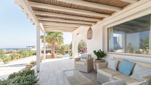 A covered outdoor seating area with wicker furniture, a glass table, and a view of the pool and palm trees. - Villa Diane (Mykonos)