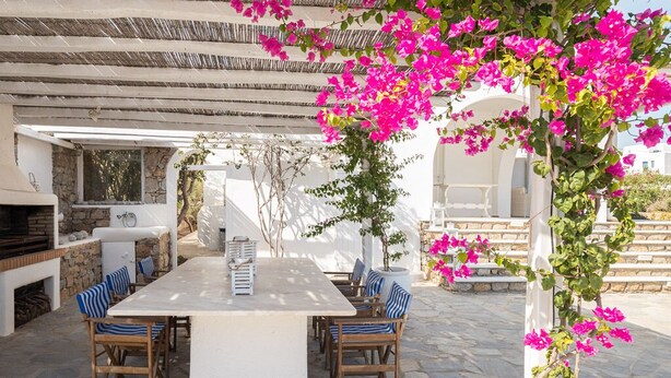 An outdoor dining area with a white table and chairs, surrounded by stone flooring and a pergola with pink bougainvillea. - Villa Diane (Mykonos)