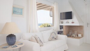 A white room with a white sofa, a table with a lamp, a TV mounted on the wall, and a window with a view of the sea. - Villa Diane (Mykonos)