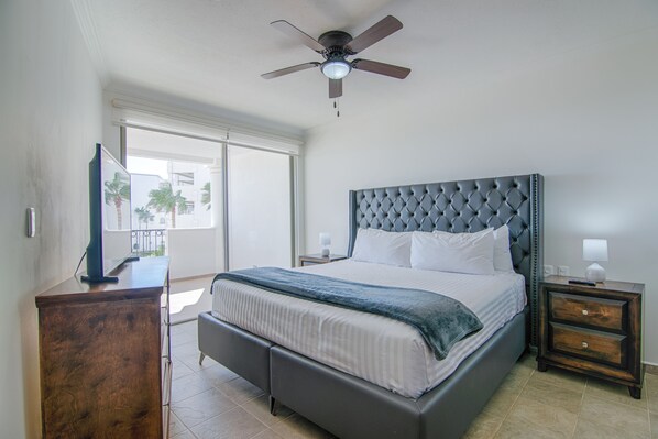 A bedroom with a large bed, a ceiling fan, a nightstand, and a view of the outdoors.