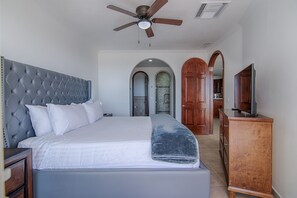 A bedroom with a tufted headboard, a large bed, a ceiling fan, and wooden doors leading to other rooms.
