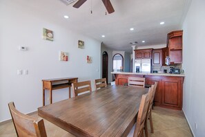 A spacious kitchen with wooden cabinets, a dining table, and chairs. There are wall-mounted pictures and a ceiling fan.