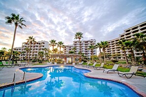 En swimmingpool omgivet af liggestole og palmer, med en hotelbygning i baggrunden.