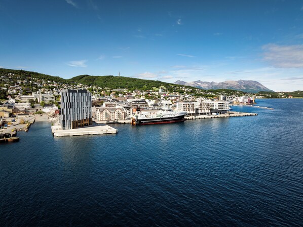Una ciudad costera con un edificio moderno imponente, un muelle con un barco, y montañas al fondo.
