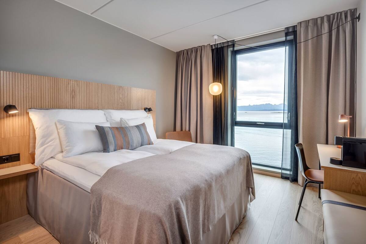 Habitación de hotel moderna con una cama grande, un escritorio, una silla, vista al mar y un cabecero de madera.