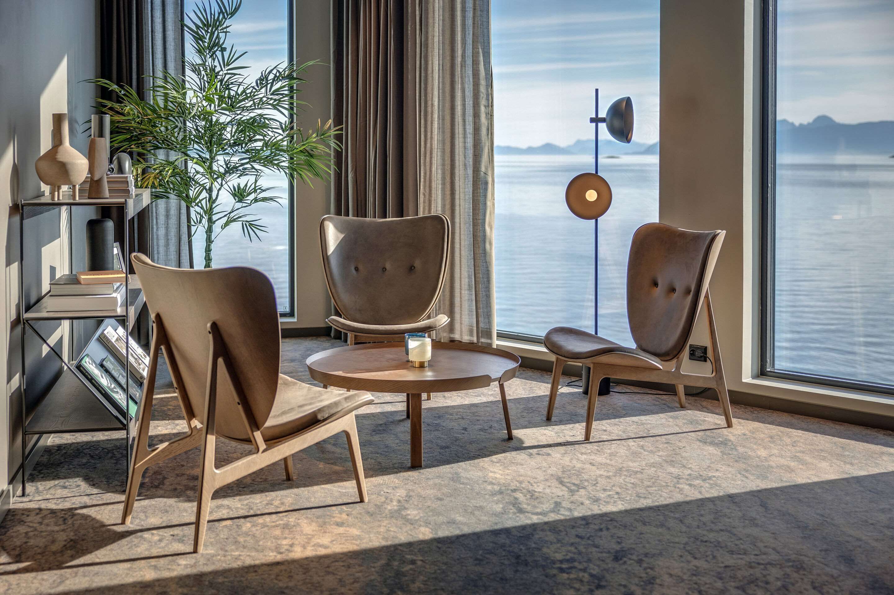 A modern room with two leather chairs, a small round table, and a bookshelf. A large window offers a view of the sea.