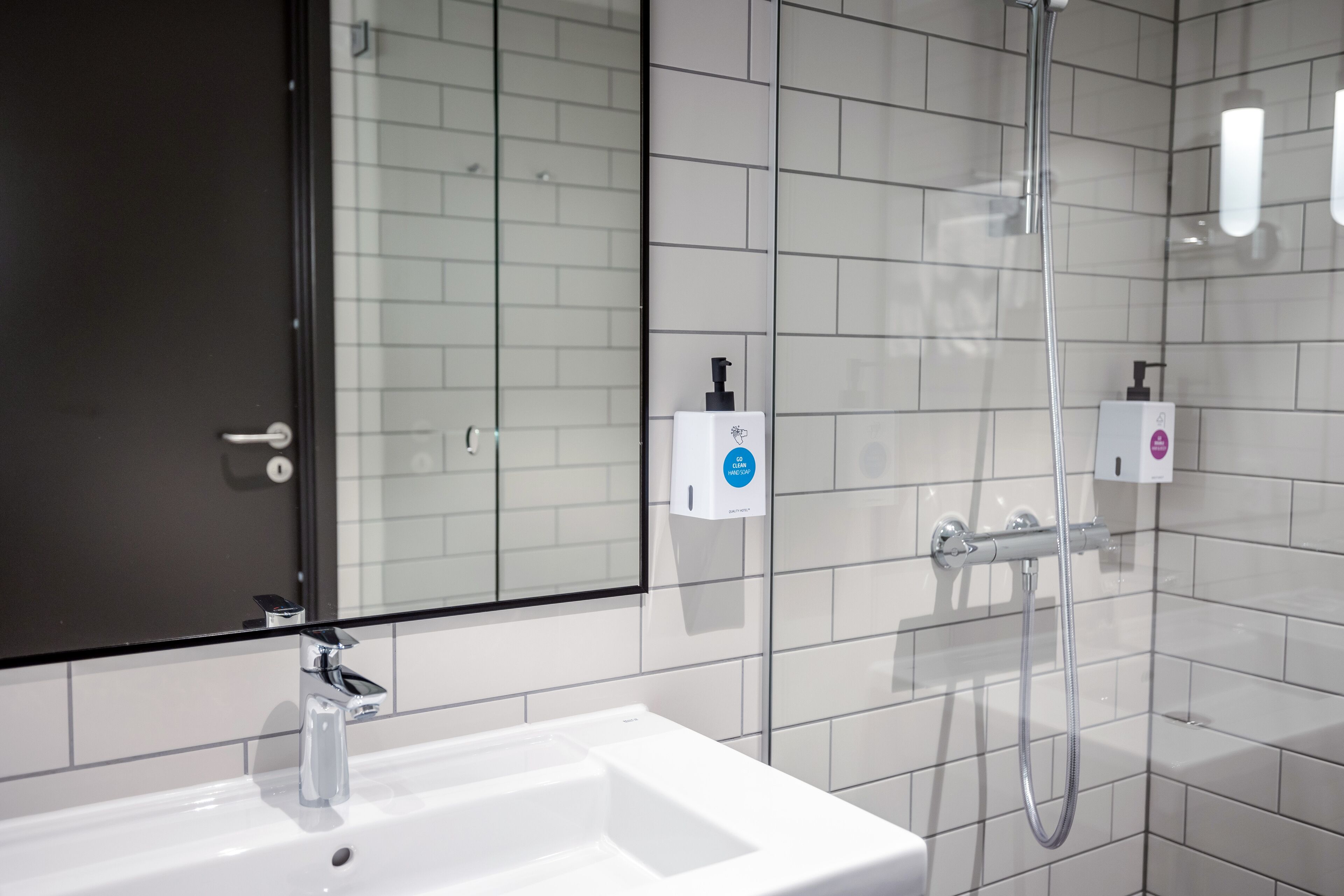 Une salle de bain moderne avec un lavabo blanc, une cabine de douche en verre et deux distributeurs de savon fixés au mur.