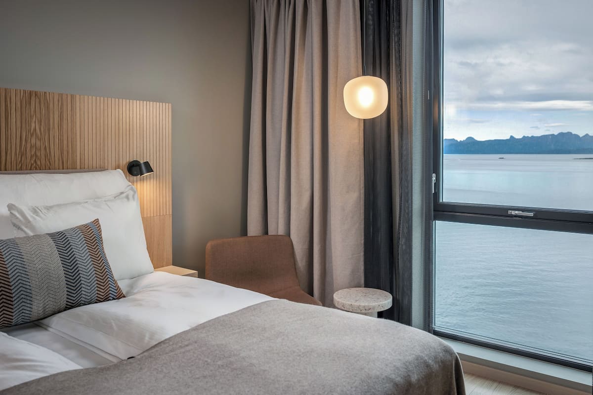 Habitación de hotel con cama, una silla, una mesita y vistas al mar a través de un ventanal grande.