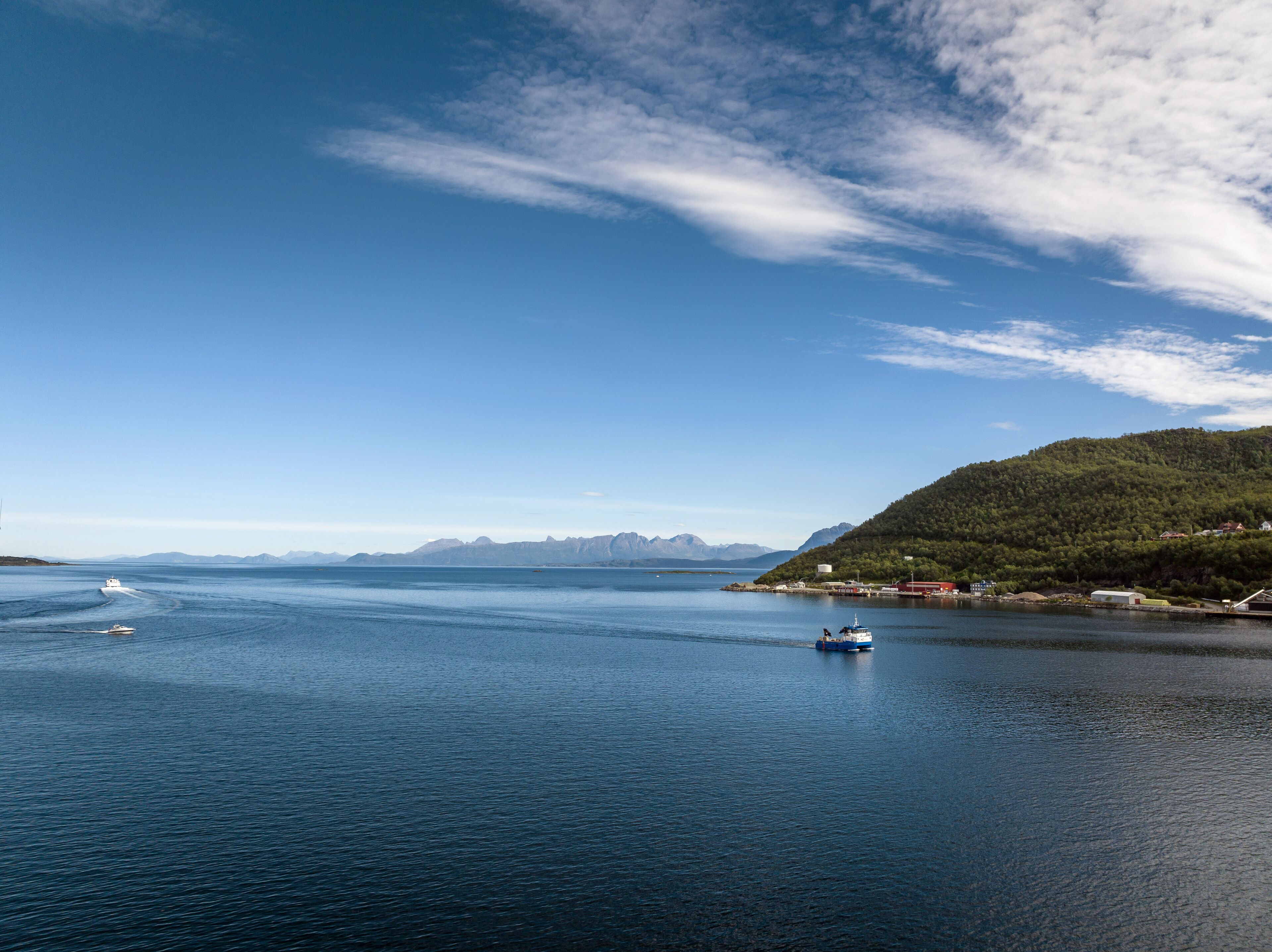 A calm sea with boats, a clear sky, and a hilly landscape in the background.