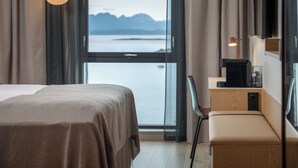 A hotel room with a bed, a desk with a coffee machine, a chair, and a large window with a view of mountains and water. - Quality Hotel Harstad (Harstad)