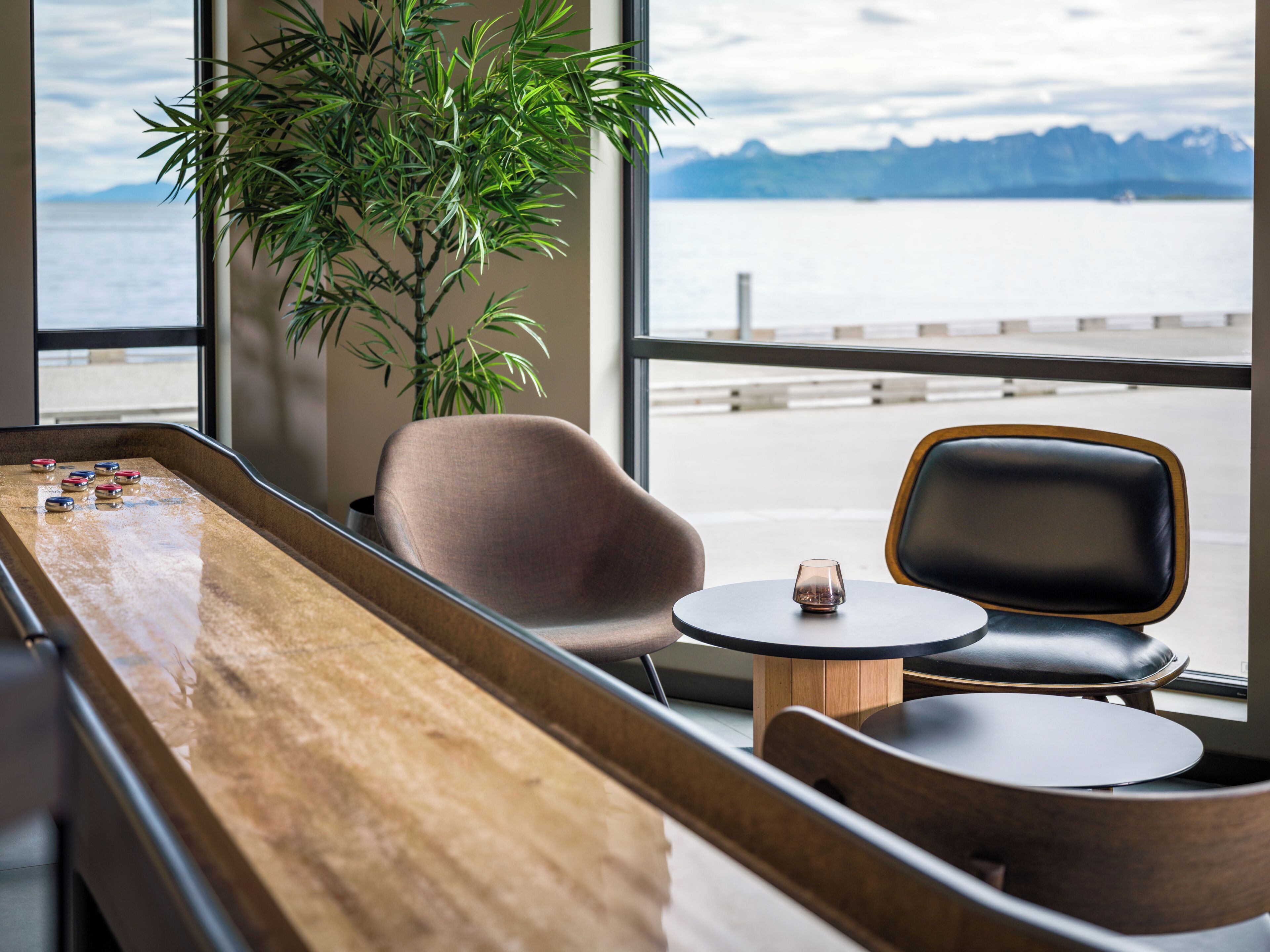 A room with a pool table, two chairs, a small table with a glass, and a large window with a view of a body of water and mountains.