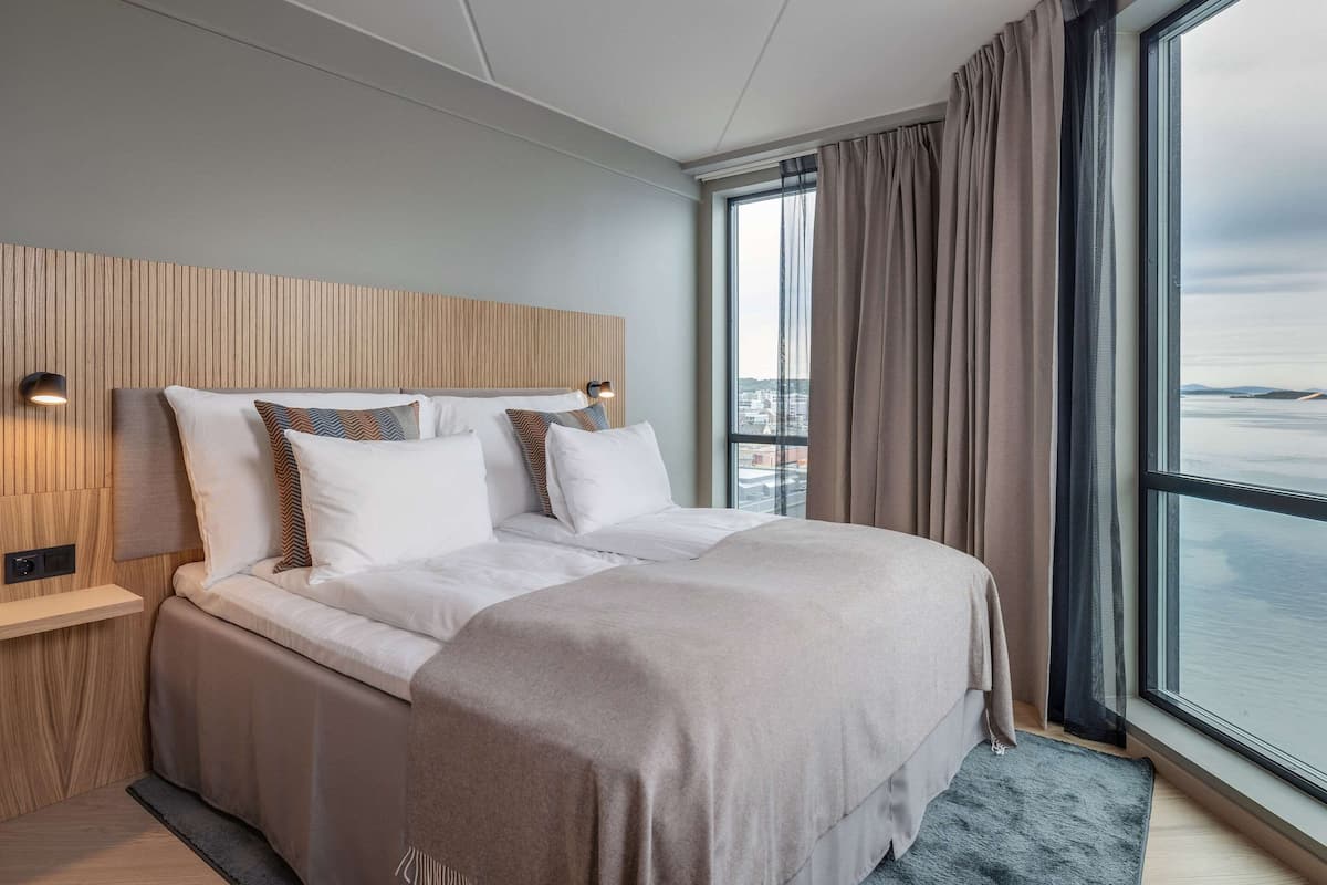 Una habitación de hotel moderna con una cama grande, cabecera de madera, mesita de noche y vistas de la ciudad a través de amplios ventanales.