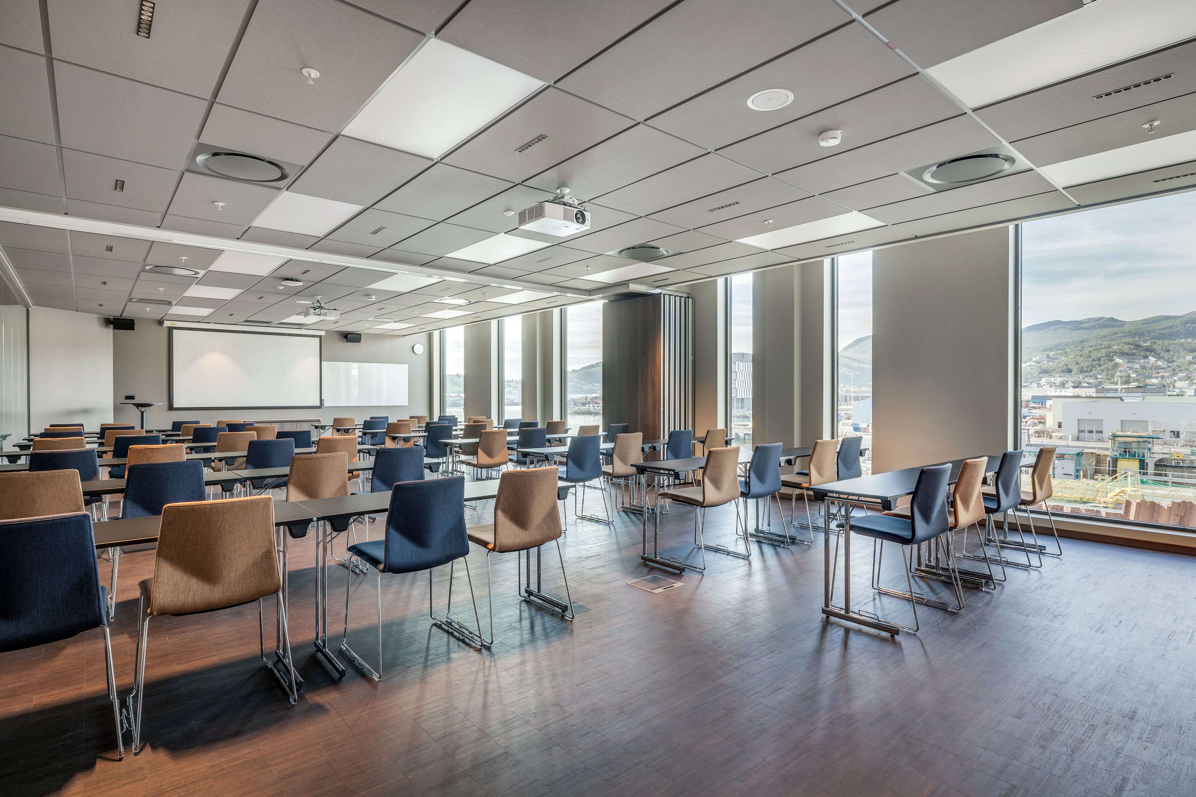 A conference room with rows of tables and chairs, large windows with a city view, and a projector screen.