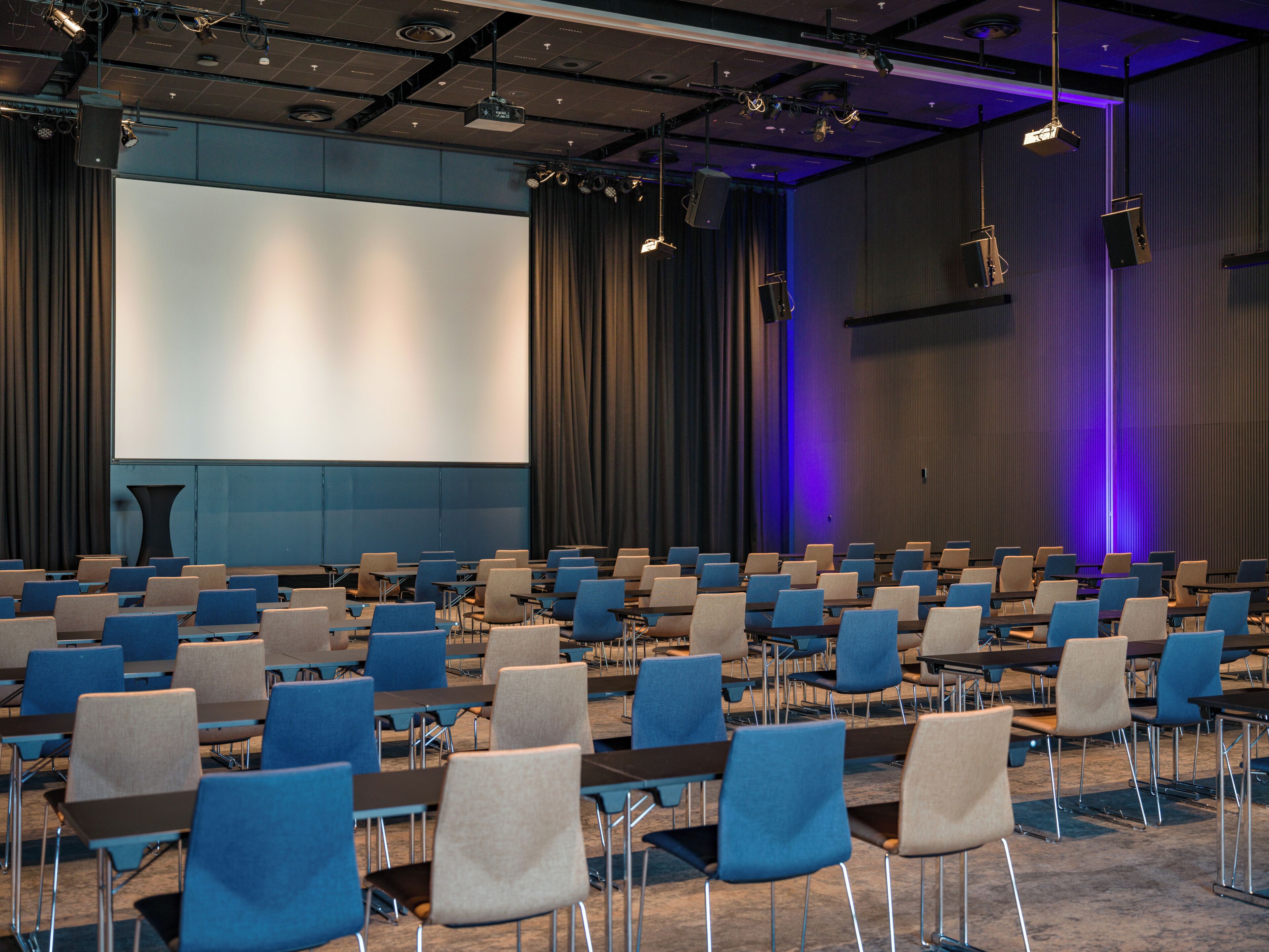 Une salle de conférence avec des rangées de chaises, un grand écran et une scène.
