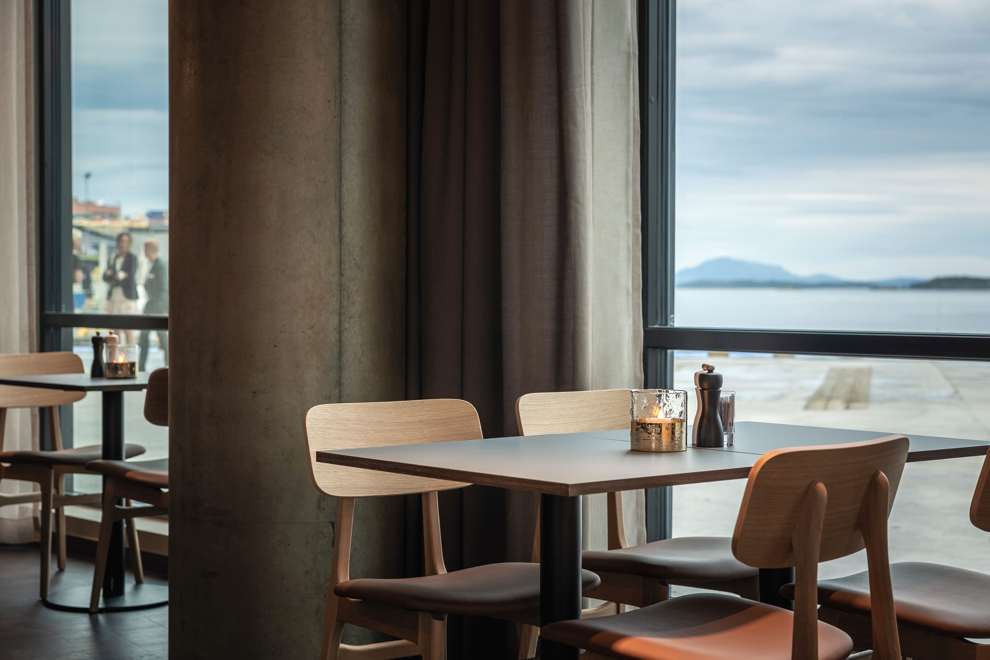 A restaurant with a view of the sea, featuring wooden tables and chairs, and a large pillar in the foreground.
