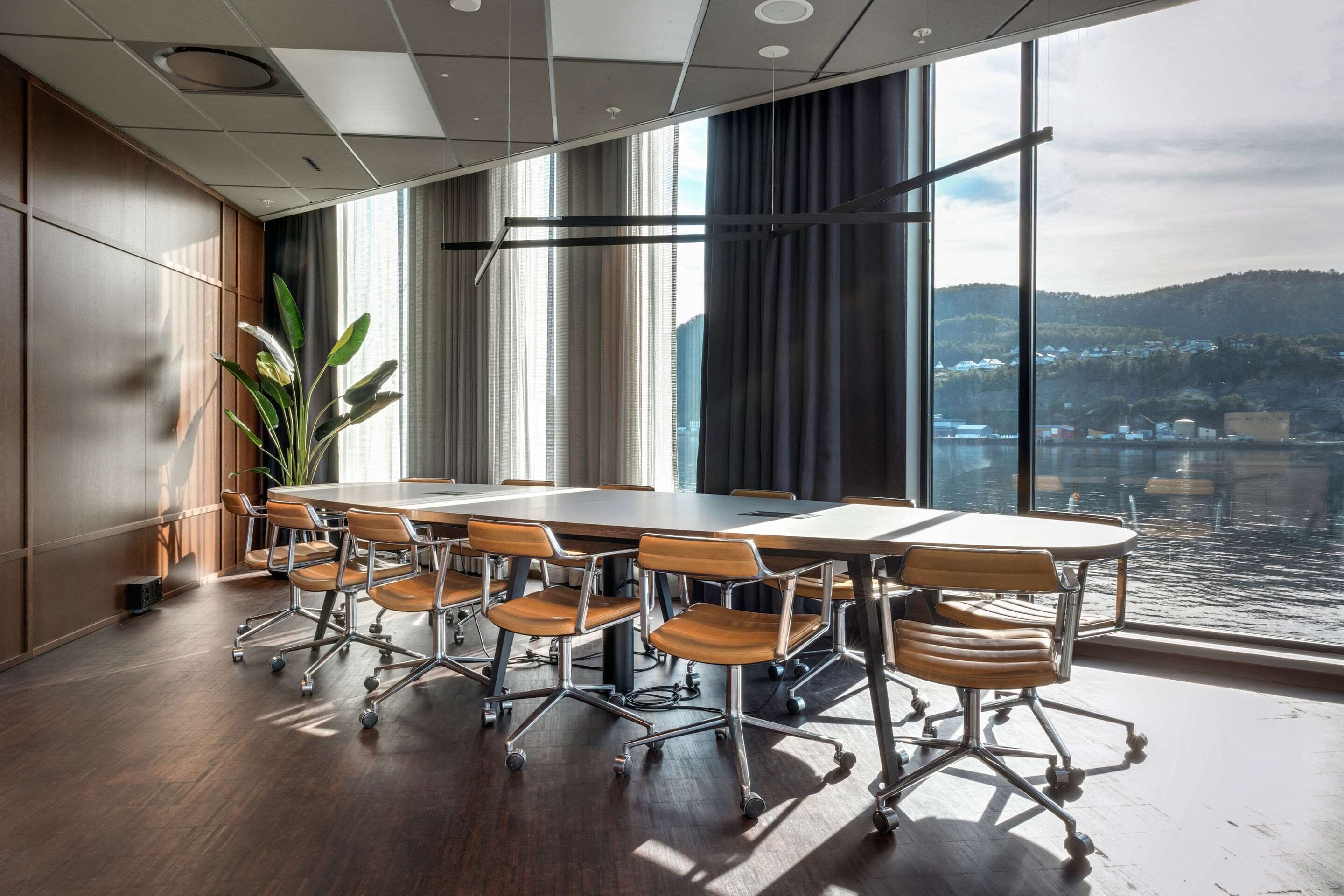 A conference room with a large table and chairs, large windows with a view of a body of water and hills, and a plant in the corner.