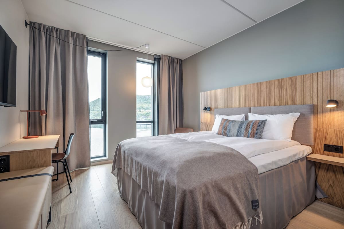 Una habitación de hotel moderna con una cama grande, un escritorio y una silla. Cuenta con una ventana con cortinas, un televisor montado en la pared y una mesita de noche con una lámpara.