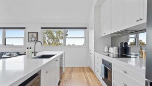 Een moderne keuken met witte kasten, een groot keukenblok met een spoelbak, en uitzicht op de omgeving door grote ramen.