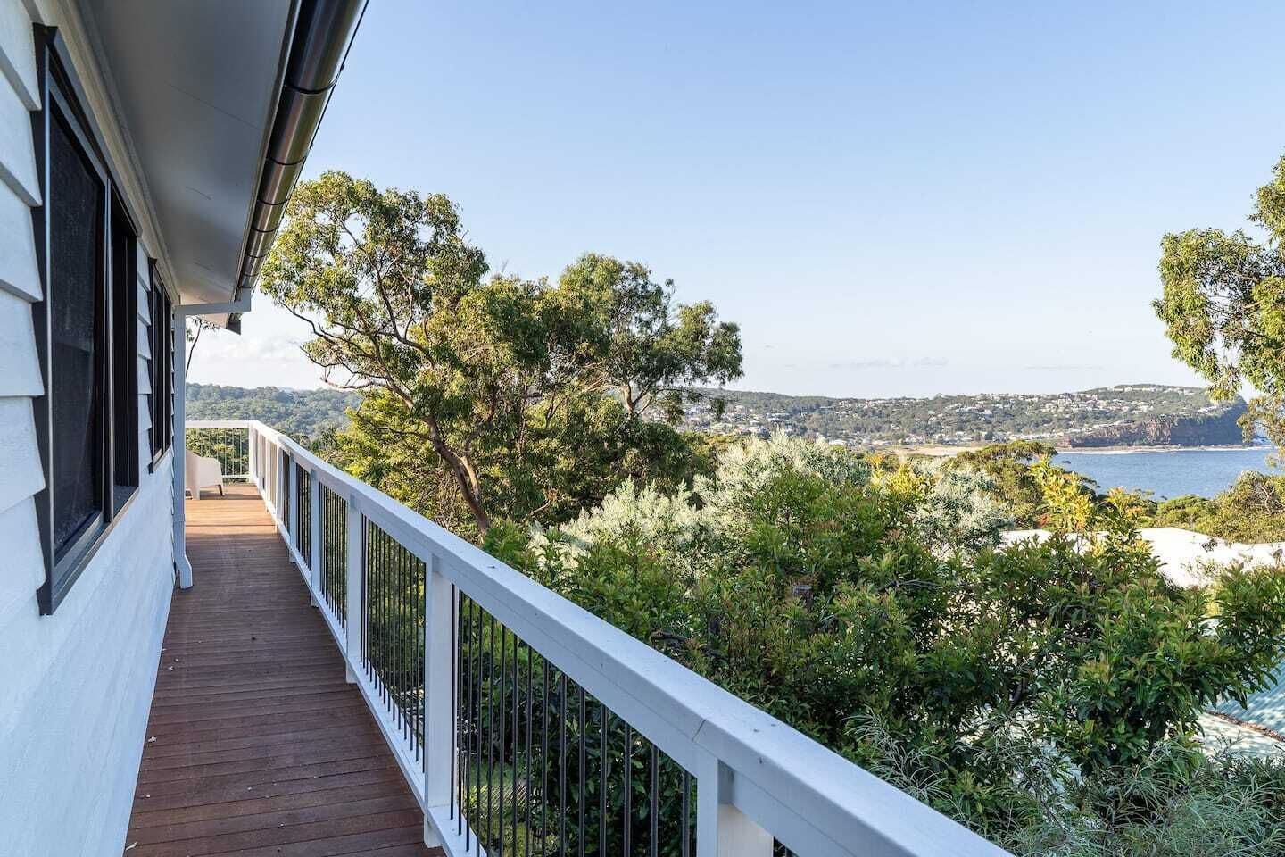 Un balcon doté d’un plancher en bois et d’une balustrade blanche, offrant une vue sur un paysage côtier avec des arbres et la mer.