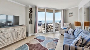 Een woonkamer met een televisie, bank en uitzicht op zee door een glazen deur.