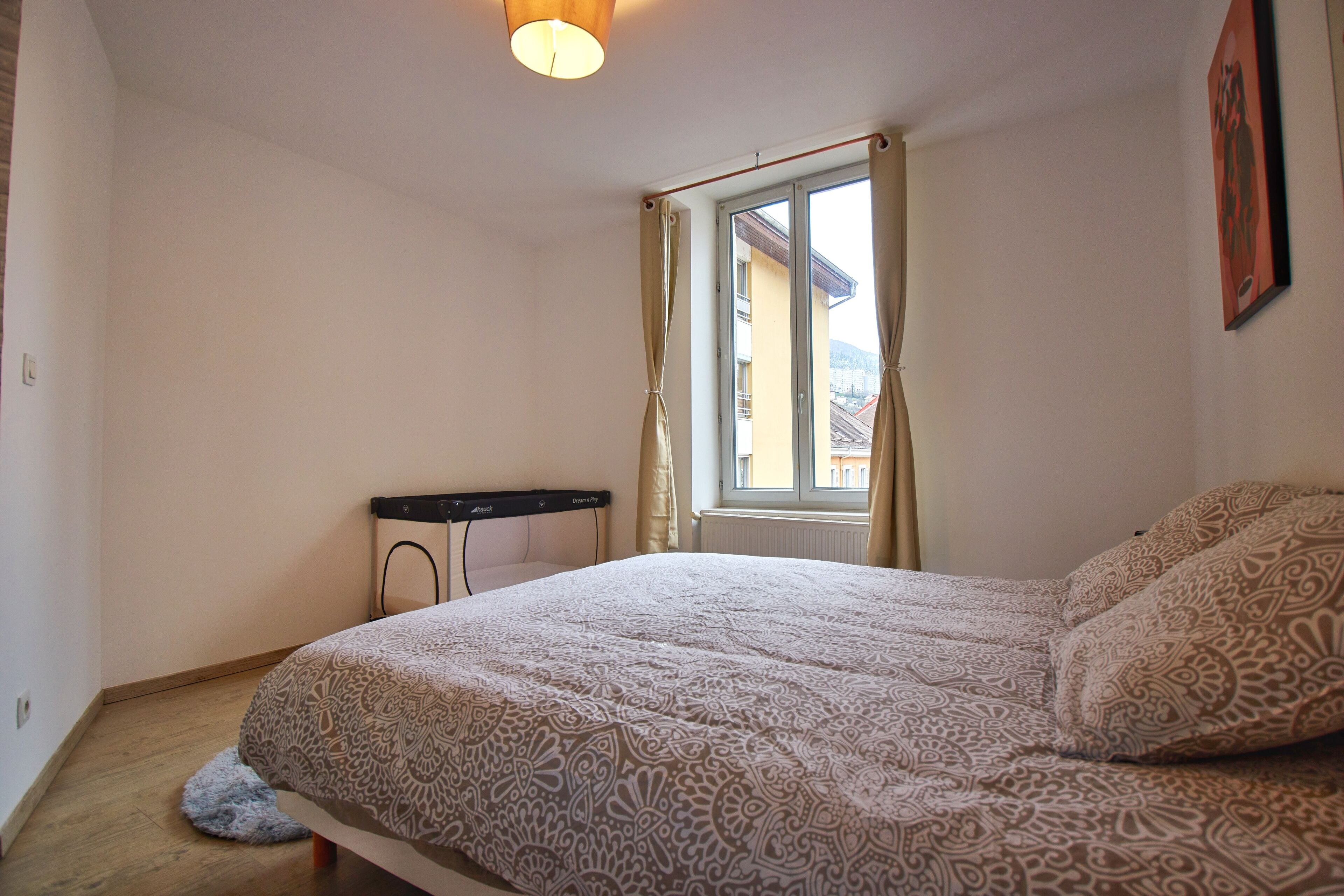 A bedroom with a bed, a desk, a window with curtains, and a radiator.