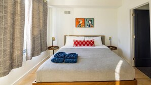 Una cama bien hecha con una colcha gris, una almohada roja y blanca, y un par de toallas azules al lado. Hay una pequeña obra de arte en la pared sobre la cama y una puerta a la derecha.