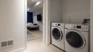 Un cuarto de lavandería con dos lavadoras de carga frontal y una cama en el dormitorio contiguo.