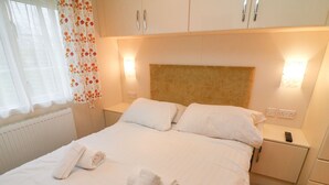 A neatly made bed with white linens and two rolled towels, a wooden headboard, bedside lamps, and a nightstand with a remote control. - The Bobbin, Number 11 (Carnforth)