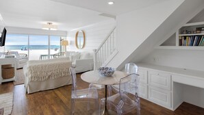 Un dormitorio amplio con una cama grande, una sala de estar con una mesa y sillas, vista a la playa y una escalera que conduce a una zona superior con una estantería.