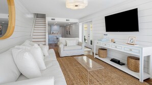 Una sala de estar moderna con un sofá blanco, una mesa de centro, un televisor sobre un mueble blanco y una escalera que lleva a una cocina.