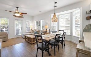 A spacious living room with a dining area, large windows with blinds, a ceiling fan, and a chandelier.