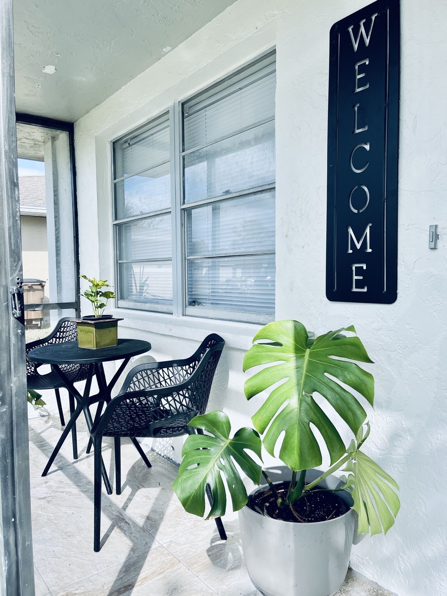 A modern outdoor seating area with a 'WELCOME' sign, large windows, and potted plants.