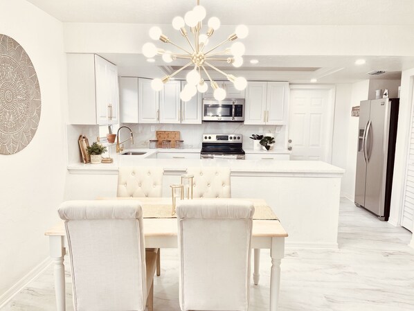 A modern kitchen with white cabinetry, a central island, a stainless steel refrigerator, and a chandelier.