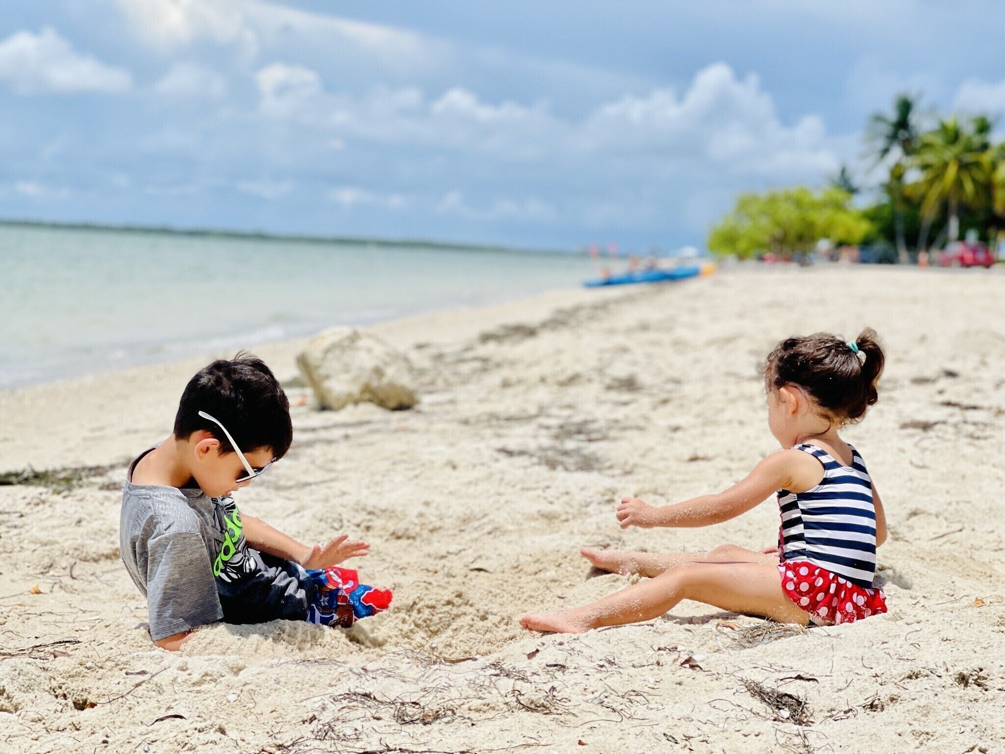 Two children playing on a sandy beach with a calm sea and palm trees in the background.